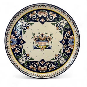 May include: A round, decorative ceramic plate with a cream-colored center and a dark blue border. The plate features floral and scroll designs in blue, brown, green, and yellow. A central floral basket design is also present.