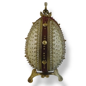 May include: A white egg-shaped decorative object with a gold and red design. The object is covered in small white beads and has a gold stand.