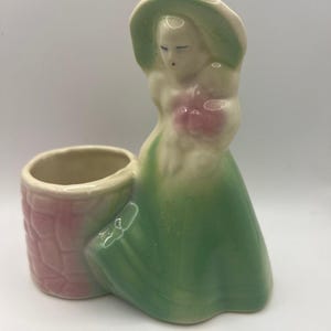 May include: A ceramic figurine planter featuring a person wearing a wide-brimmed green hat and a long green dress, holding a pink bouquet. Next to the figure is a cylindrical container with a textured pink brick-patterned exterior.
