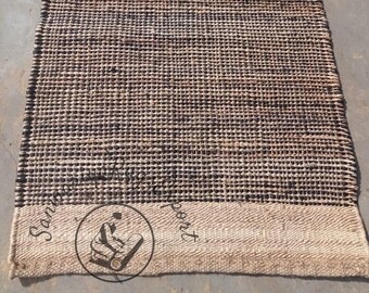 May include: A brown and black woven rug with a white border. The rug has a textured surface and is made of natural fibers. The rug is approximately 2.5 feet wide and 8 feet long.