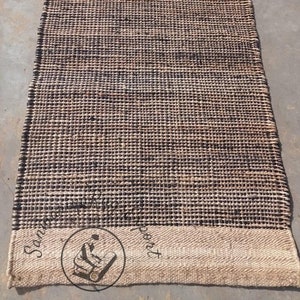 May include: A brown and black woven rug with a white border. The rug has a textured surface and is made of natural fibers. The rug is approximately 2.5 feet wide and 8 feet long.