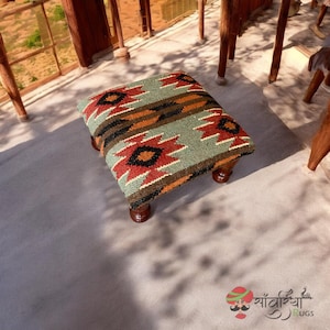 May include: A small, square footstool with a patterned top. The fabric features a geometric design in red, black, orange, and brown on a green background. The footstool has four short, dark wooden legs.