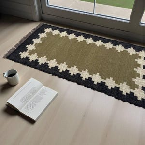May include: A rectangular woven rug featuring an olive green center, a black border, and a cream-colored geometric cross pattern. The rug has short fringe on the ends. Beside the rug sits a white ceramic mug filled with dark liquid and an open book.