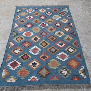 May include: A rectangular blue rug with a geometric diamond pattern in shades of red, orange, yellow, and white. The rug has a fringed edge and is suitable for home decor.