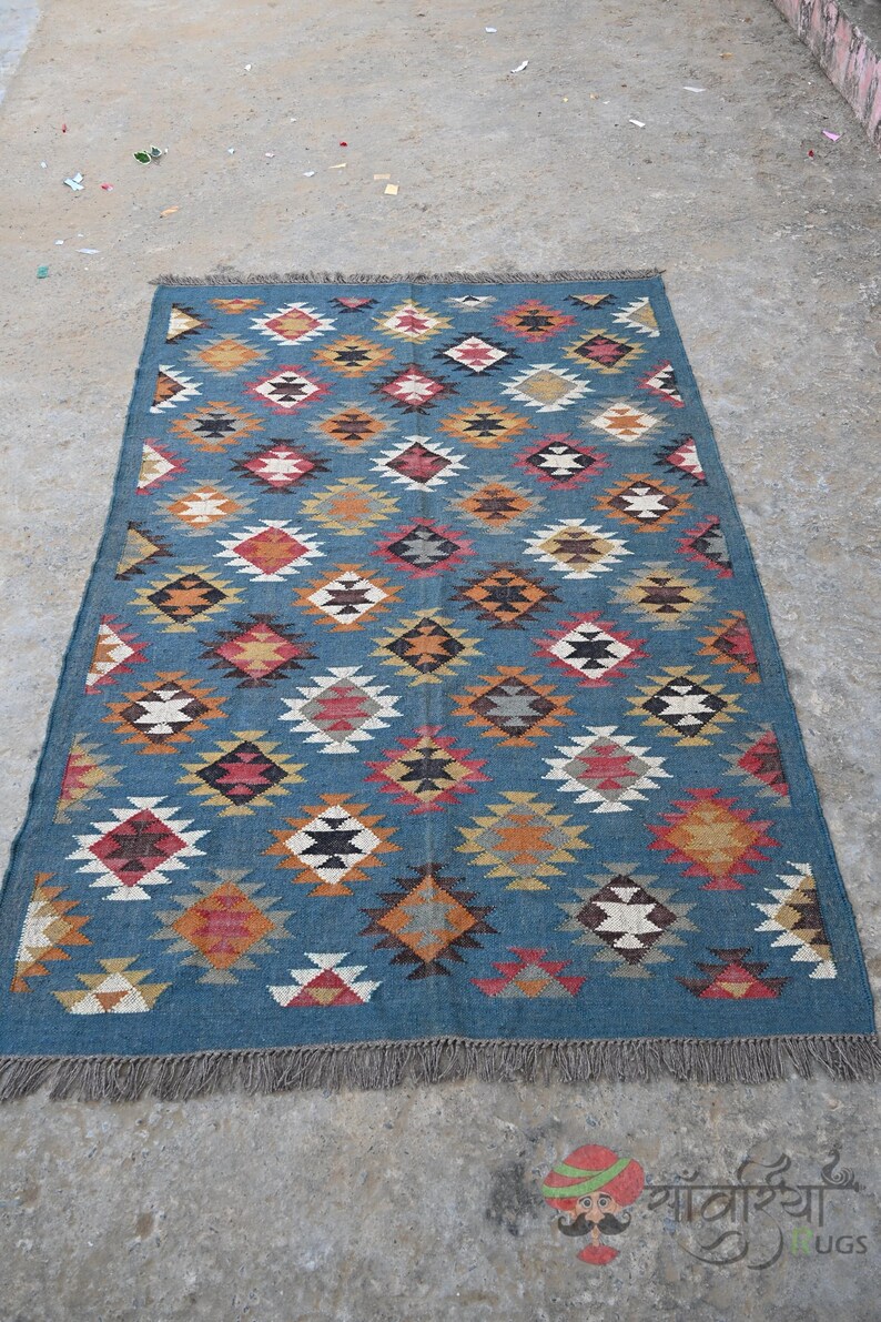 May include: A rectangular blue rug with a geometric diamond pattern in shades of red, orange, yellow, and white. The rug has a fringed edge and is suitable for home decor.