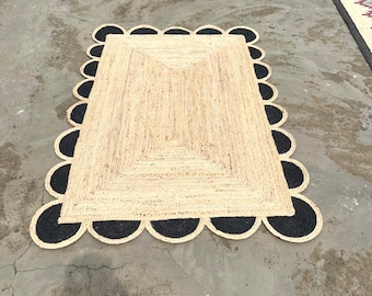 May include: A rectangular area rug made of natural tan woven jute fibers. The rug features a central rectangular pattern and is bordered by a series of solid black circular scalloped edges that extend outward along all four sides of the rug.