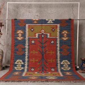 May include: A rectangular rug with a central red field, bordered by blue and accented with geometric patterns in white, gold, and brown. The rug is hanging from a metal bar, showcasing its intricate design and fringed edges. The rug is likely made of natural fibers.