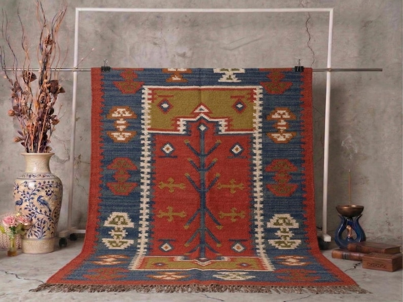 May include: A rectangular rug with a central red field, bordered by blue and accented with geometric patterns in white, gold, and brown. The rug is hanging from a metal bar, showcasing its intricate design and fringed edges. The rug is likely made of natural fibers.