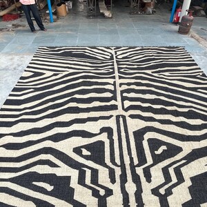 A large black and off-white zebra-patterned rug with fringe lies flat on a concrete floor, with industrial equipment in the background.