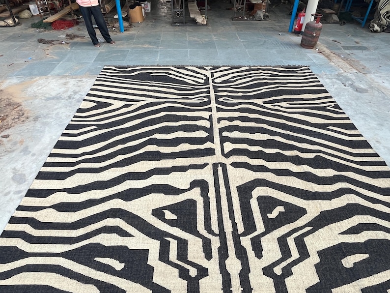 A large black and off-white zebra-patterned rug with fringe lies flat on a concrete floor, with industrial equipment in the background.