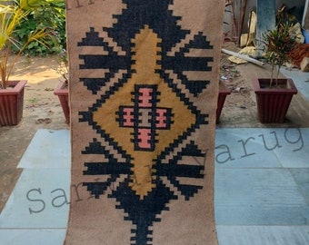 May include: A rectangular beige rug with a geometric pattern in black, gold, and pink. The central design features a diamond shape with a cross-like element. The rug has a fringed edge and is suitable for home decor.