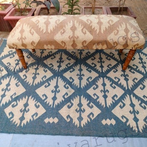 Può includere: Una panca con motivo marrone e beige con gambe in legno si trova su un tappeto con motivo blu e beige. Il tappeto ha un design geometrico.