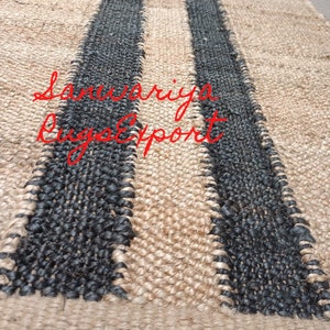 May include: A natural fiber rug with two black stripes woven into the beige material. The rug is made of jute and has a natural, earthy look. The text "Sanwariya Rug Export" is visible in the lower left corner of the image.