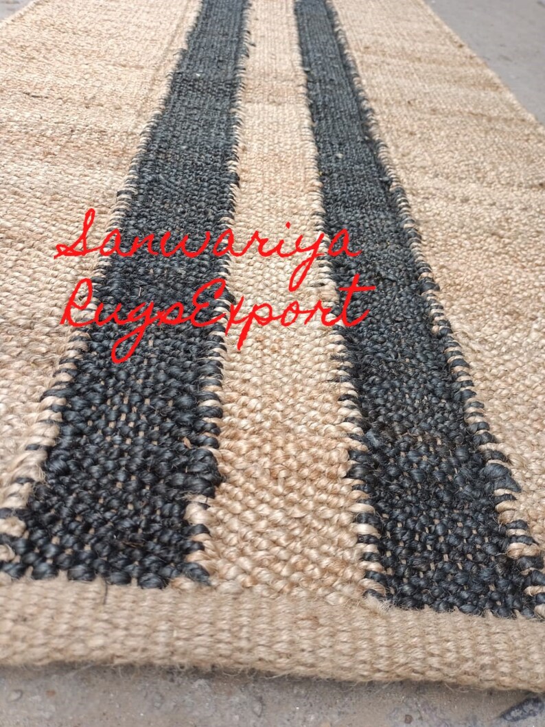 May include: A natural fiber rug with two black stripes woven into the beige material. The rug is made of jute and has a natural, earthy look. The text "Sanwariya Rug Export" is visible in the lower left corner of the image.