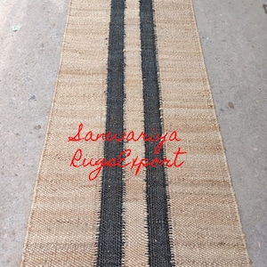 May include: A natural fiber rug with two black stripes running down the center. The rug is made of jute and has a natural, earthy look. The text "Sanwariya Rugs Export" is visible in the lower center of the rug.