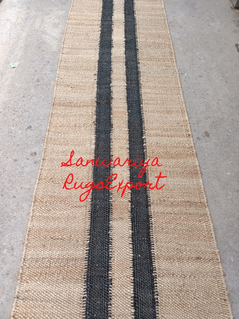 May include: A natural fiber rug with two black stripes running down the center. The rug is made of jute and has a natural, earthy look. The text "Sanwariya Rugs Export" is visible in the lower center of the rug.