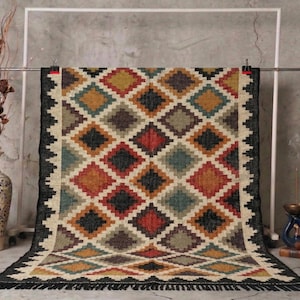 May include: A rectangular rug with a geometric diamond pattern in shades of red, black, beige, yellow, and teal. The rug has a black border with fringe. The rug is displayed against a gray background.