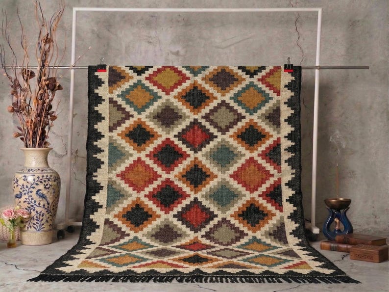 May include: A rectangular rug with a geometric diamond pattern in shades of red, black, beige, yellow, and teal. The rug has a black border with fringe. The rug is displayed against a gray background.
