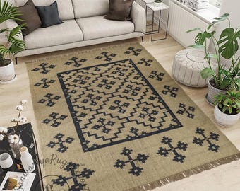May include: A rectangular area rug featuring a tan base colour with a dark charcoal geometric pattern consisting of repeating cross-like shapes and a central border. The rug has a fringed edge on the top and bottom sides and a visible watermark.