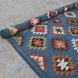 May include: A rolled-up blue rug with a geometric pattern. The rug features repeating diamond and square shapes in shades of red, white, orange, and black. The rug is partially unrolled, revealing the pattern.