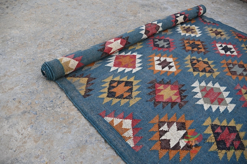 May include: A rolled-up blue rug with a geometric pattern. The rug features repeating diamond and square shapes in shades of red, white, orange, and black. The rug is partially unrolled, revealing the pattern.
