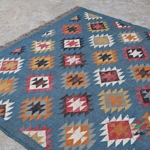 May include: A blue rug with a repeating geometric pattern of squares in various colors, including orange, red, white, and black. The rug has a fringed edge and is likely made of woven fabric.