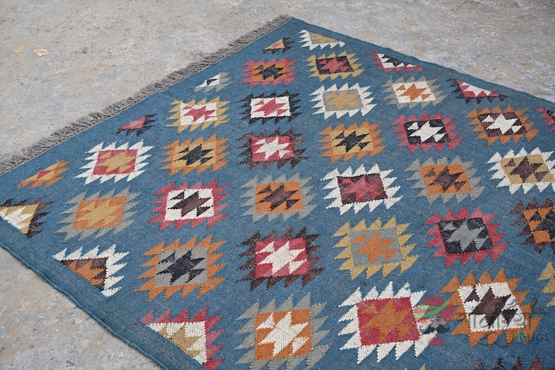 May include: A blue rug with a repeating geometric pattern of squares in various colors, including orange, red, white, and black. The rug has a fringed edge and is likely made of woven fabric.
