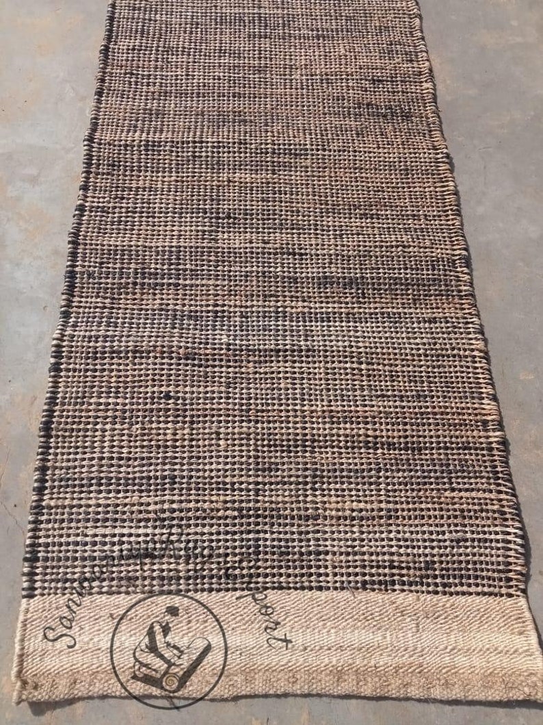 May include: A brown and black woven rug with a white border. The rug has a textured surface and is made of natural fibers. The rug is approximately 2.5 feet wide and 8 feet long.