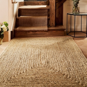 May include: A natural jute rug with a geometric design, laid on a wooden floor. The rug is beige and has a textured, woven appearance. A staircase with brown carpet and wooden accents is in the background. The rug measures approximately 200cm x 140cm.