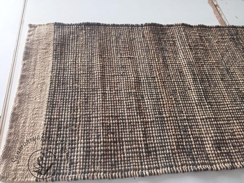 May include: A woven rug with a natural jute border and a dark brown center. The rug is made of natural fibers and has a textured surface.