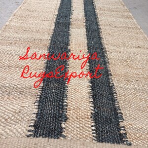 May include: A natural fiber rug with two black stripes running down the center. The rug is made of jute and has a natural, earthy look. The text "Sanwariya Rug Export" is visible in the lower center of the rug.