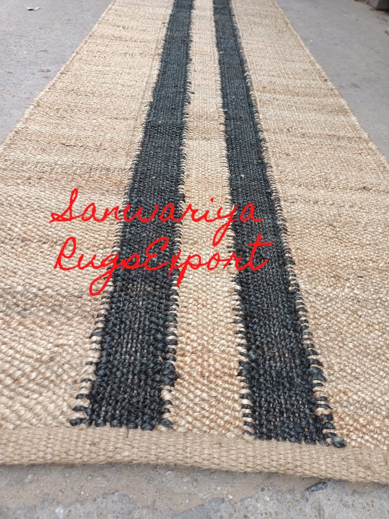 May include: A natural fiber rug with two black stripes running down the center. The rug is made of jute and has a natural, earthy look. The text "Sanwariya Rug Export" is visible in the lower center of the rug.