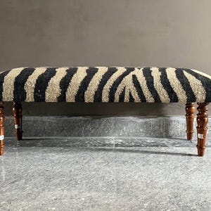 May include: A rectangular bench with a zebra stripe pattern in black and beige. The bench has four turned wooden legs in a dark brown finish. The bench is a decorative furniture piece.