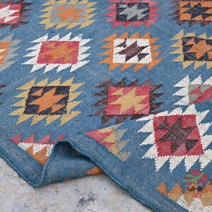 May include: A blue rug with a repeating geometric pattern. The pattern features squares with star-like designs in shades of orange, red, brown, and white. The rug's border is a darker shade of blue, and the rug is folded over.