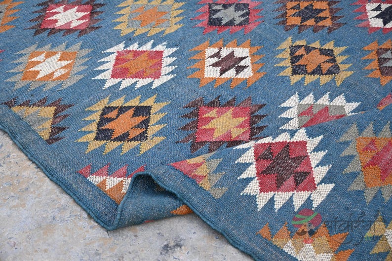 May include: A blue rug with a repeating geometric pattern. The pattern features squares with star-like designs in shades of orange, red, brown, and white. The rug's border is a darker shade of blue, and the rug is folded over.