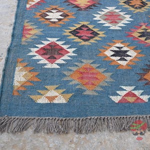 May include: A blue woven rug with a geometric pattern in shades of red, orange, white, and black. The rug has a fringe along the bottom edge and is likely made of natural fibers.