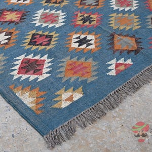 May include: A blue rug with a geometric pattern of squares in various colors, including white, orange, and red. The rug has a fringe along one edge and is likely made of woven material.