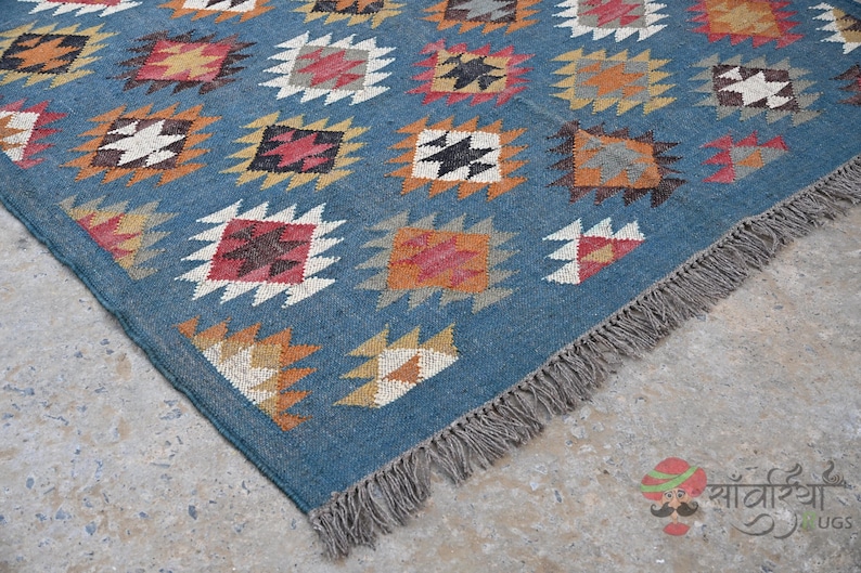 May include: A blue rug with a geometric pattern of squares in various colors, including white, orange, and red. The rug has a fringe along one edge and is likely made of woven material.