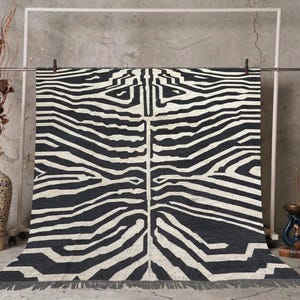 May include: A black and white zebra-striped rug with a fringed edge. The rug is hanging from a metal bar against a gray concrete wall. A decorative vase with dried branches and a blue and white vase are in the background.