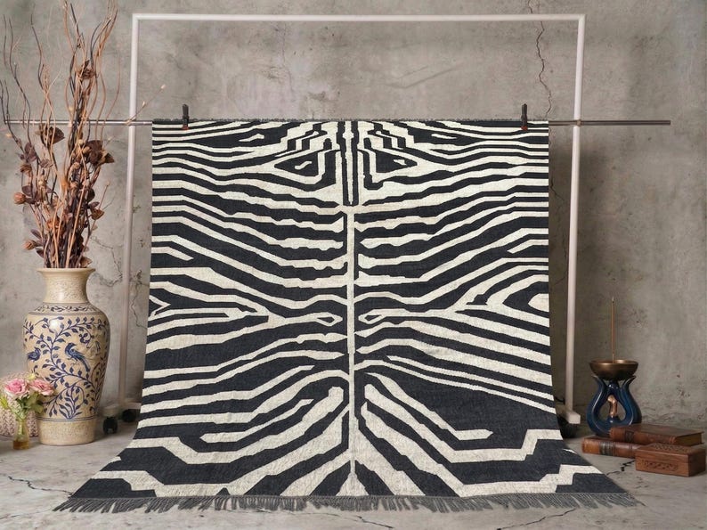 May include: A black and white zebra-striped rug with a fringed edge. The rug is hanging from a metal bar against a gray concrete wall. A decorative vase with dried branches and a blue and white vase are in the background.