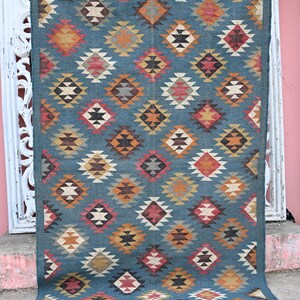 May include: A rectangular, teal-colored rug with a repeating diamond pattern in shades of orange, red, yellow, and white. The rug has a fringed edge and is displayed against a white, ornate background.