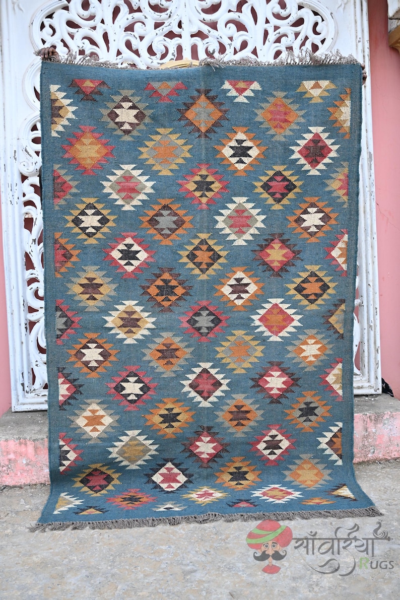 May include: A rectangular, teal-colored rug with a repeating diamond pattern in shades of orange, red, yellow, and white. The rug has a fringed edge and is displayed against a white, ornate background.