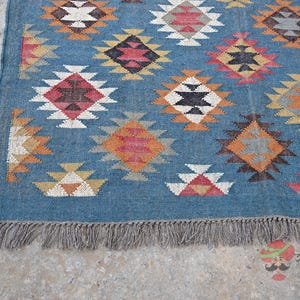 May include: A blue rug with a geometric pattern in shades of red, white, black, and orange. The rug has a fringe along the bottom edge. The pattern consists of diamond shapes with smaller triangles inside.