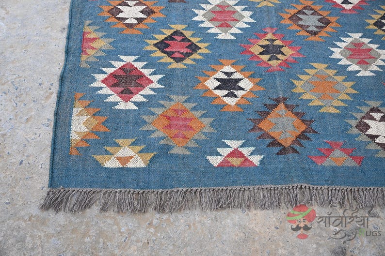 May include: A blue rug with a geometric pattern in shades of red, white, black, and orange. The rug has a fringe along the bottom edge. The pattern consists of diamond shapes with smaller triangles inside.