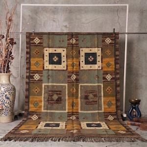 May include: A rectangular area rug with a geometric pattern in shades of brown, gold, and green. The rug features square and diamond shapes, with a fringed edge. The rug is displayed on a metal frame against a neutral background.
