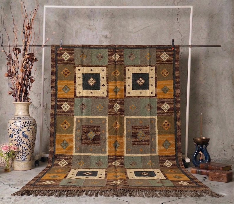 May include: A rectangular area rug with a geometric pattern in shades of brown, gold, and green. The rug features square and diamond shapes, with a fringed edge. The rug is displayed on a metal frame against a neutral background.