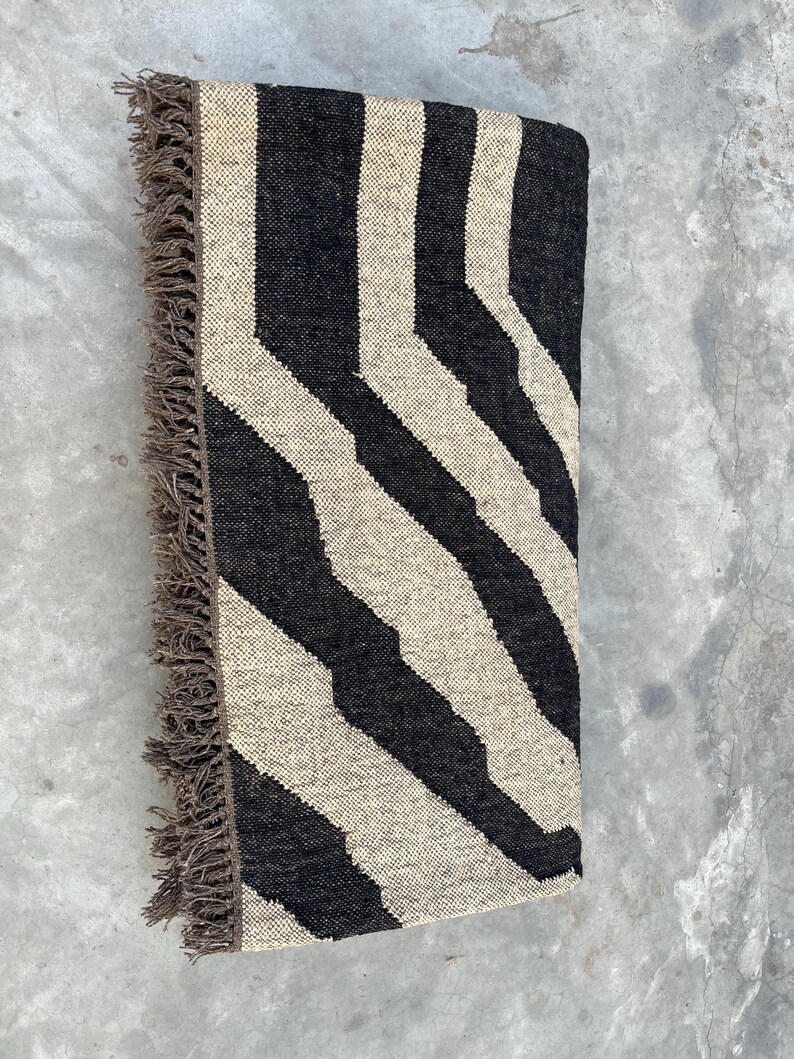 A black and off-white zebra-patterned rug with fringe is folded in half, lying on a concrete surface.