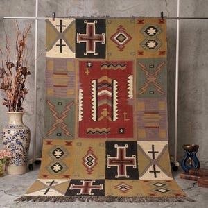May include: A rectangular, patterned rug with a patchwork design. The rug features geometric shapes, crosses, and diamond patterns in shades of red, brown, green, and beige. It has a fringed edge.
