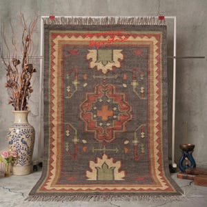 May include: A rectangular, dark gray rug with a central geometric design in red, green, and beige. The rug has a fringed edge and a border with a repeating pattern. The rug is displayed on a white rack.