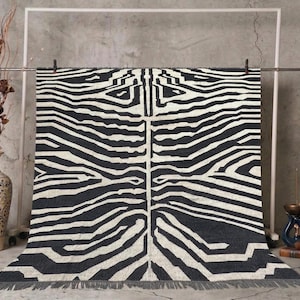 A black and off-white zebra-patterned rug with fringe is hung from a white metal frame, with a vase of dried plants and books on either side.